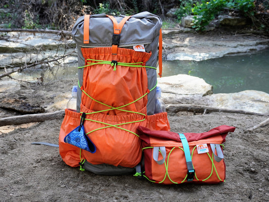 Orange and gray backpack and hip pack on a natural outdoor setting with water and rocks in the background.