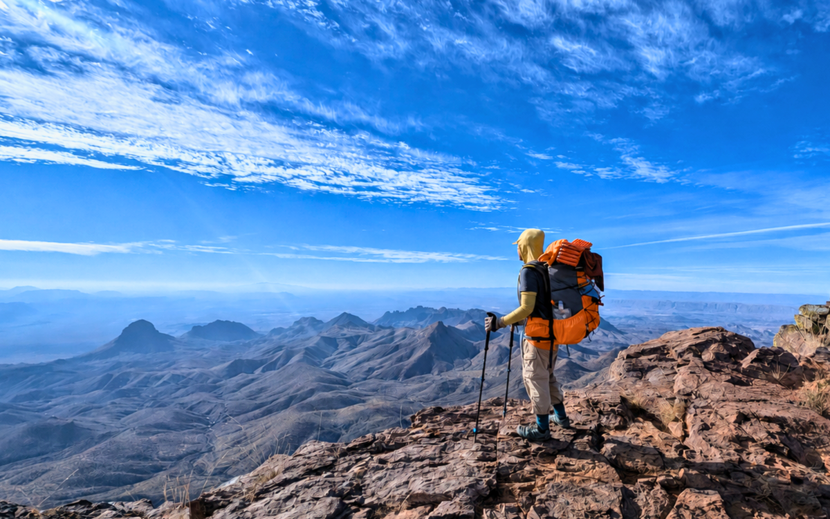 Sojurnr founder Jennifer Villamin wearing the Resolute 50-55 women’s backpack and holding trekking poles on a rocky peak in Big Bend National Park, Texas, overlooking a vast mountain range under a bright blue sky with wispy clouds.
